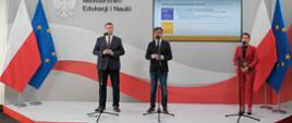 Widok z przodu - na podwyższeniu stoją minister Czarnek, mężczyzna w czarnym garniturze i kobieta ubrana na czerwono, za nimi flaga Polski.