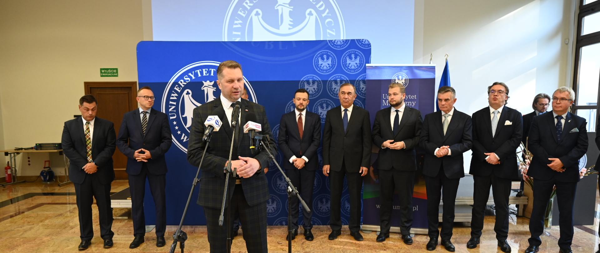 Na dużej oświetlonej przez wpadające przez duże okna światło sali pod niebieską ścianką stoi w szeregu kilku mężczyzn w garniturach, przed nimi minister Czarnek stoi i mówi do kilku mikrofonów na stojakach.