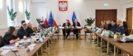 Widok z przodu na niewielka salę, przy ustawionych w prostokąt stołach siedzą ludzie, u szczytu stołu siedzi minister Czarnek i mężczyzna w garniturze, za nimi flagi Polski i UE, dalej na ścianie godło, na ścianie po lewej dwa okna.