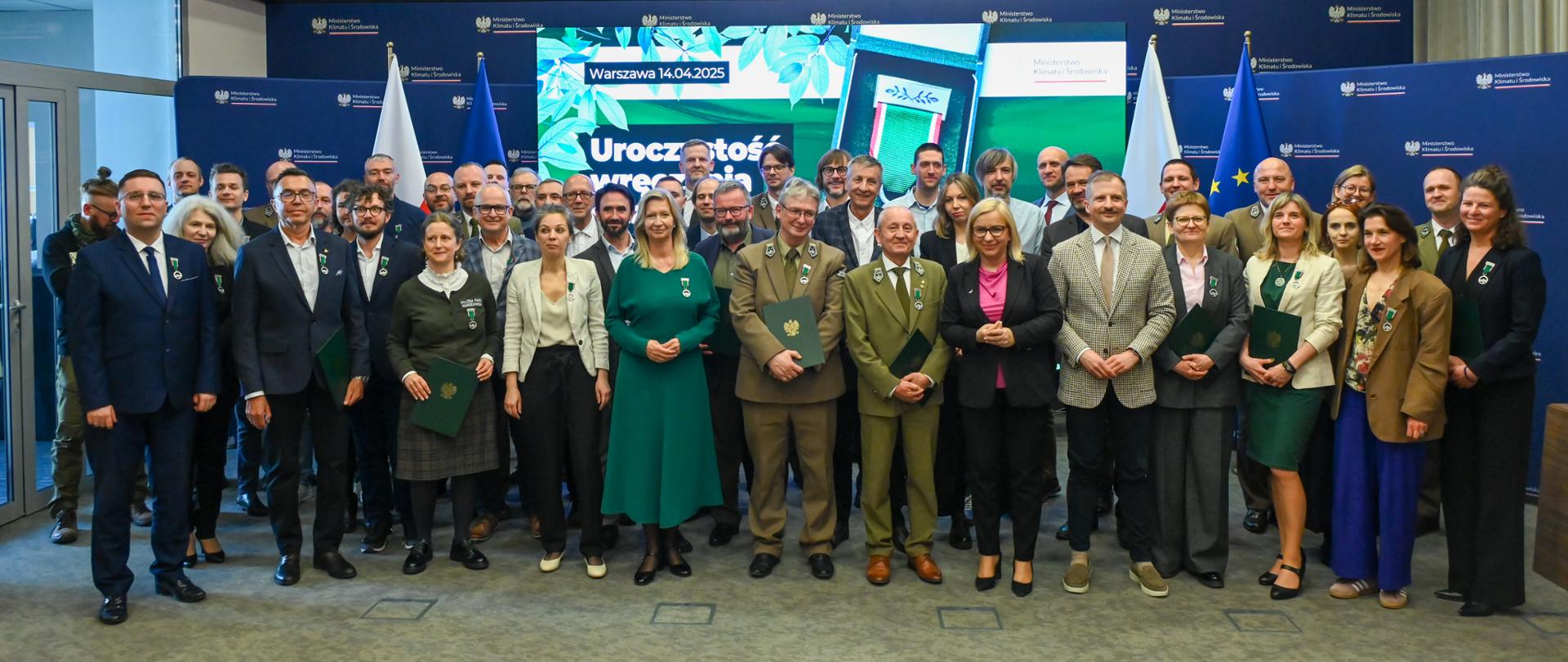 Zdjęcie grupowe z uroczystości wręczenia Odznak Honorowych za Zasługi dla Ochrony Środowiska i Klimatu przedstawia postaci osób odznaczonych, ministry Pauliny Hennig-Kloski, wiceministra Mikołaja Dorożały oraz w tle wśród gości wiceministra Krzysztofa Bolesty.
