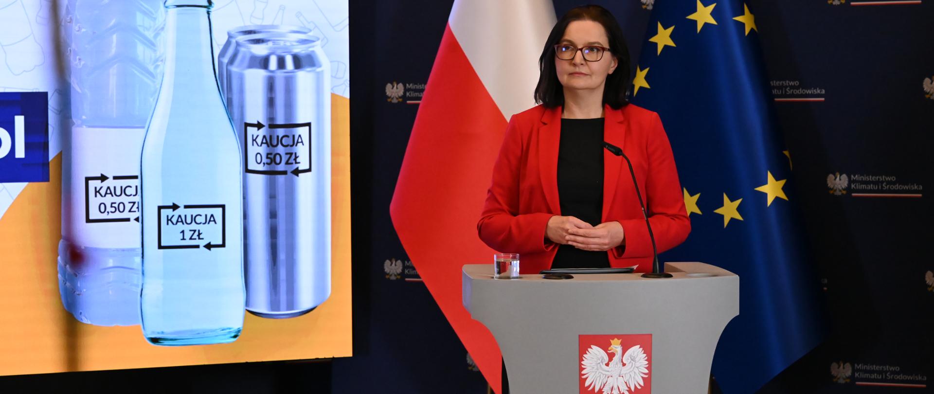 Wiceministra klimatu i środowiska Anita Sowińska podczas konferencji prasowej. Stoi za mównicą. Za nią widoczny jest fragment prezentacji, na której widzimy butelki ze znakiem kaucji, a po prawej stronie zdjęcia widoczne są dwie flagi - flaga Polski i Unii Europejskiej. 