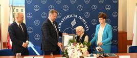 Za drewnianym stołem stoi minister Czarnek, mężczyzna w garniturze i kobieta w niebieskiej marynarce, minister podaje siedzącej przed nim kobiecie w czarnej kamizelce dokument w ramkach.