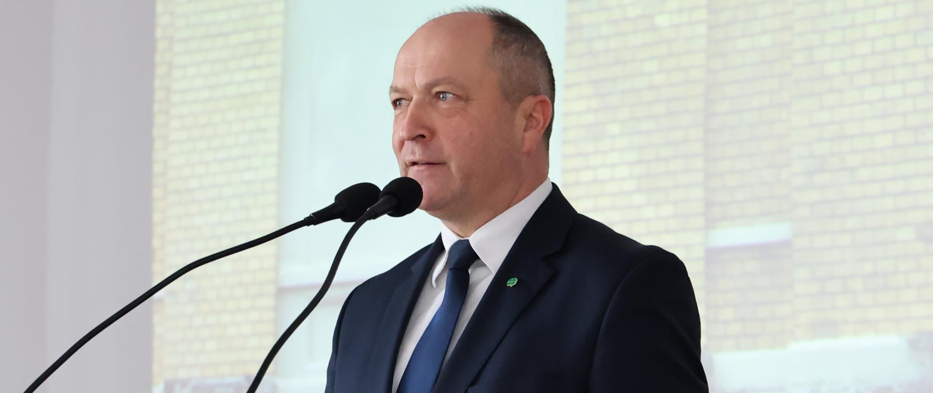Minister Henryk Kiepura, sekretarz stanu w MEN, w pozycji wyprostowanej przemawia do dwóch mikrofonów, znajdując się przy mównicy. Minister ma na sobie granatowy garnitur, białą koszulę i granatowy krawat. W klapie wpięta jest czterolistna koniczyna. W tle wyświetlana jest prezentacja multimedialna.