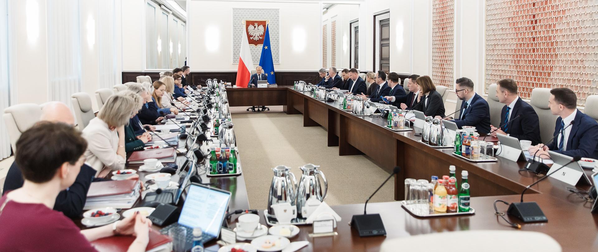 Posiedzenie Rady Ministrow. Ministrowie wraz z Premierem siedzą w dużej jasnej sali przy dużych, drewnianych stołach.