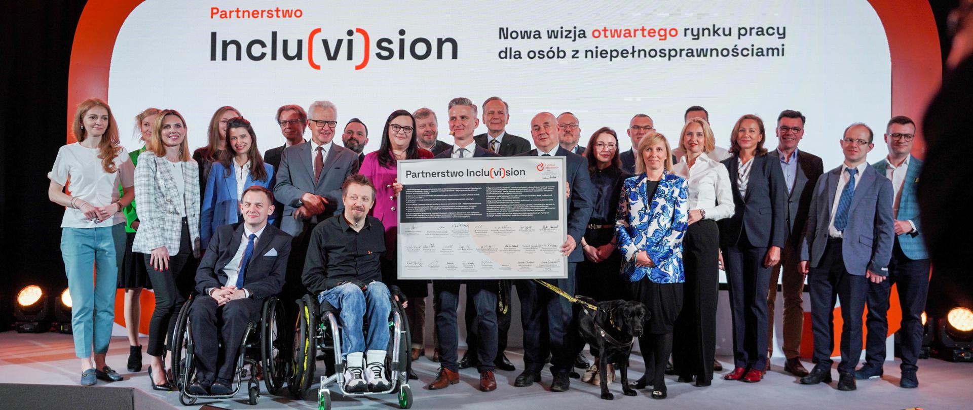 Grupa osób na scenie. Mężczyzna trzyma duży baner z napisem "Partnerstwo Incluvision". Obok niego stoi wiceminister Małgorzata Jarosińska-Jedynak.