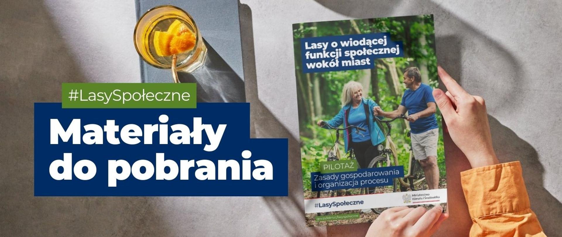 Na zdjęciu widnieje szary notes, na którym stoi pomarańczowa szklanka z wodą. Obok w dłoniach trzymana jest książka pt. Lasy o wiodącej funkcji społecznej wokół miast. Na zdjęciu jest napis: #LasySpołeczne, Materiały do pobrania.