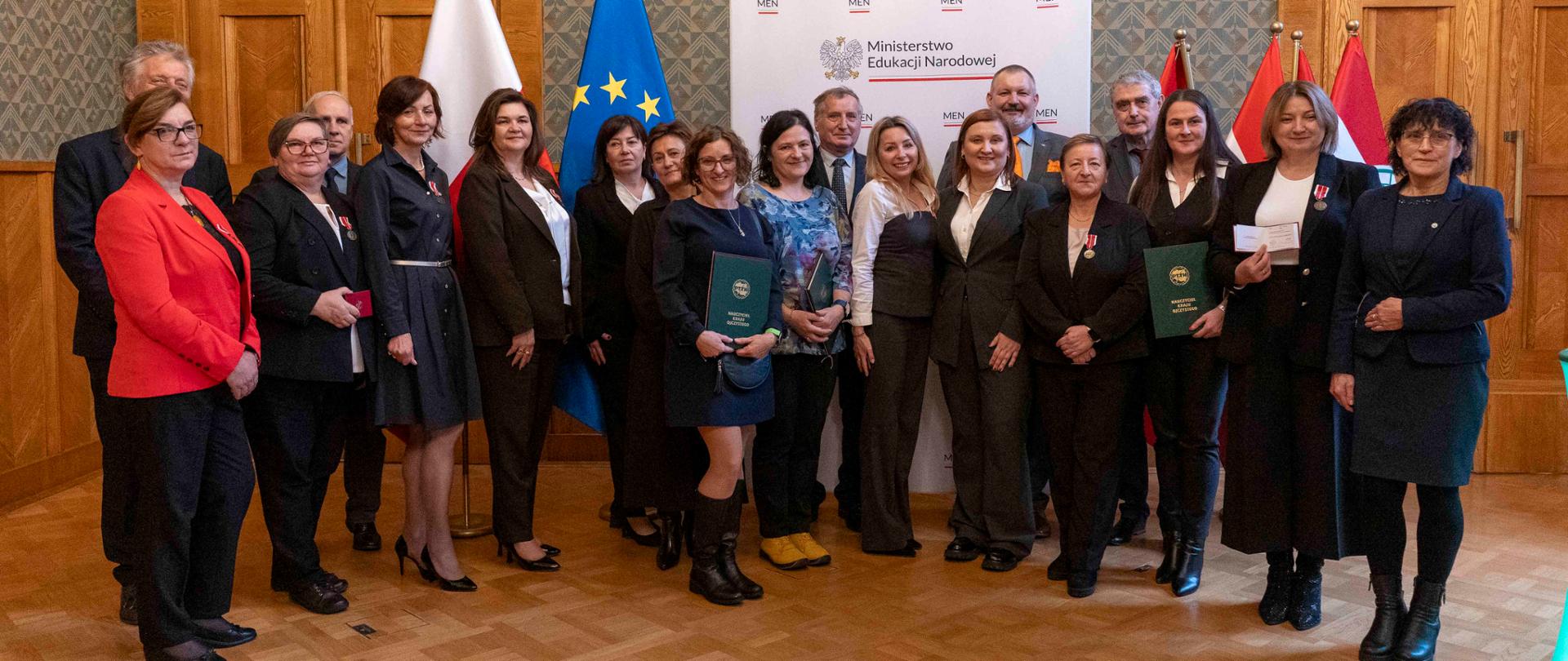 Zdjęcie grupowe kilkunastu osób (nagrodzonych i organizatorów) stojących razem w sali. W tle widoczna jest ścianka z logo Ministerstwa Edukacji Narodowej oraz flagi Polski, Unii Europejskiej i PTTK. Osoby trzymają dyplomy i Medale Komisji Edukacji Narodowej.