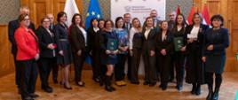 Zdjęcie grupowe kilkunastu osób (nagrodzonych i organizatorów) stojących razem w sali. W tle widoczna jest ścianka z logo Ministerstwa Edukacji Narodowej oraz flagi Polski, Unii Europejskiej i PTTK. Osoby trzymają dyplomy i Medale Komisji Edukacji Narodowej.