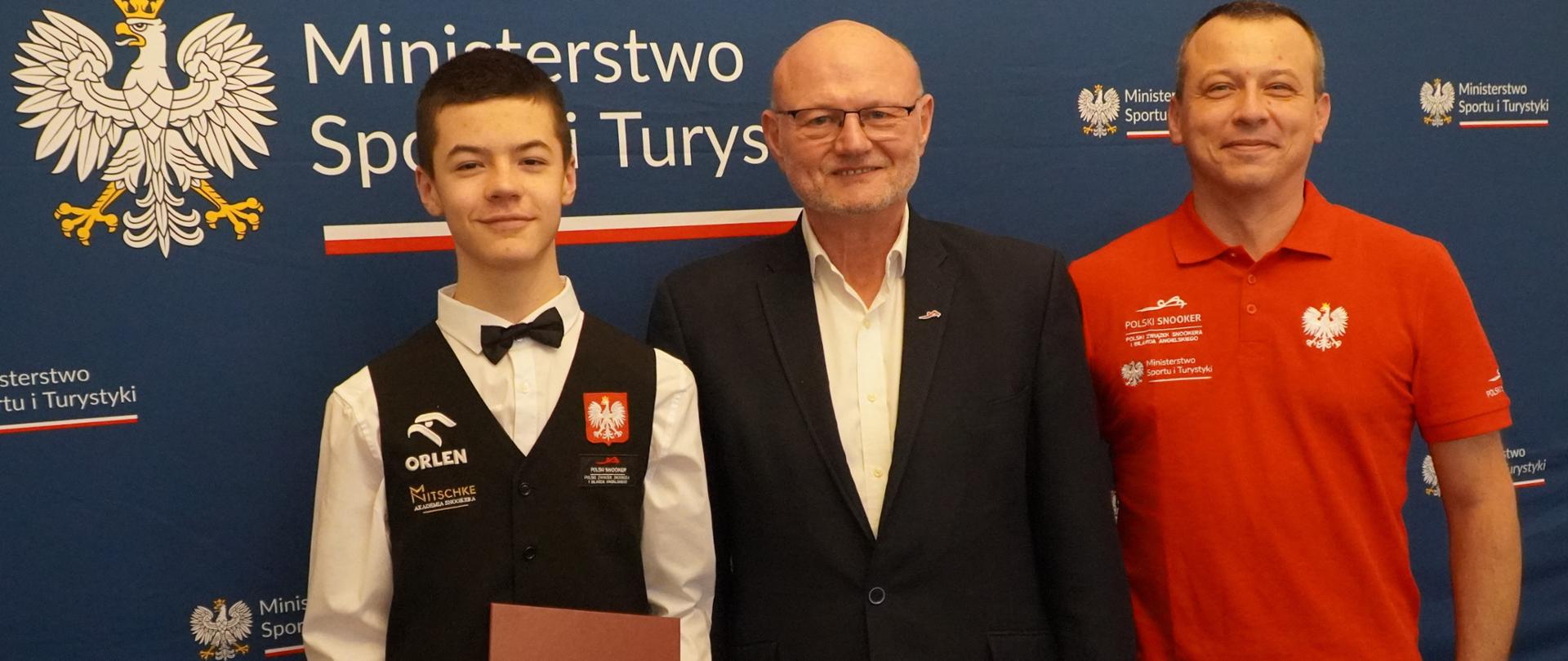 Od lewej stoją: Michał Szubarczyk, podsekretarz Stanu w Ministerstwie Sportu i Turystyki Ireneusz Nalazek i Kamil Szubarczyk (tata i trener Michała Szubarczyka) na tle granatowej ścianki z logotypami MSiT. Michał Szubarczyk trzyma w ręku teczkę z napisem "Ministerstwo Sportu i Turystyki".