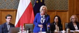 Wiceminister Machałek w niebieskim ubraniu stoi za stołem, za nią ściana w zielony wzorek i flagi Polski i UE.