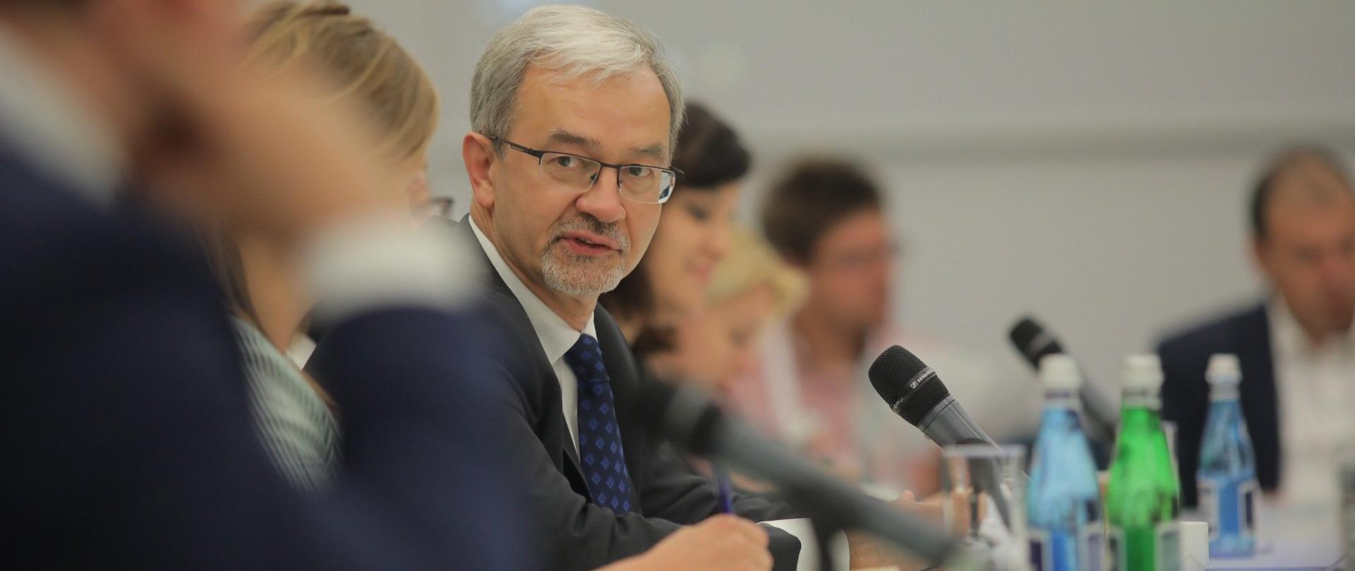 minister Jerzy Kwieciński mówi do mikrofonu stojącego na stole i jest zwrócony do swojego rozmówcy