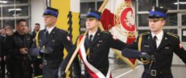 Trzech mężczyzn w czarnych galowych mundurach niesie sztandar.