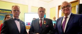 W sali z żółto-białymi ścianami stoi w rzędzie trzech mężczyzn w garniturach.