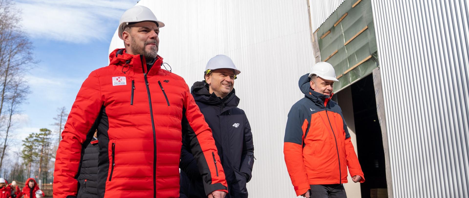 Zdjęcie przedstawia Ministra Sportu i Turystyki Sławomira Nitrasa spacerującego obok dużej hali. Minister jest ubrany w czarną kurtkę i kask budowlany, a razem z nim spacerują osoby w czerwonych kurtkach z naklejką COS Zakopane oraz również ubrane w kaski budowlane. W tle oprócz hali widoczne są drzewa.