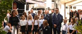 Na trawie przed budynkiem stoi grupa ubranych na galowo dzieci i kilkoro dorosłych.