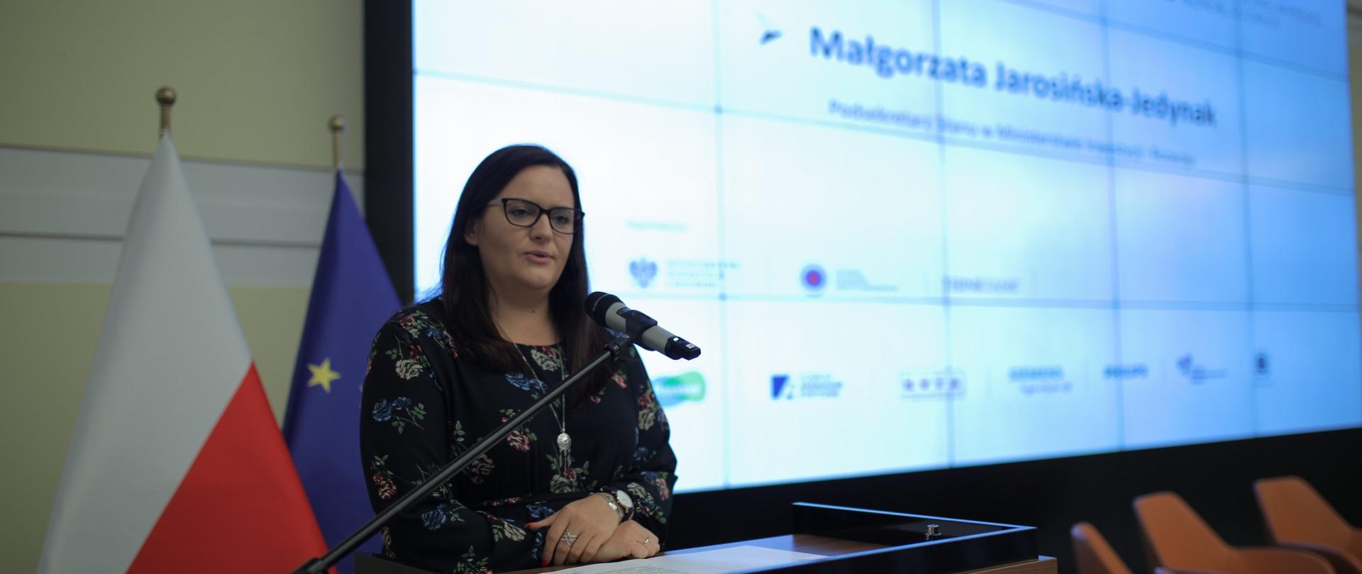 Wiceminister Małgorzata Jarosińska-Jedynak na scenie przy mównicy podczas przemówienia, z tyłu flagi: Polski i Unii Europejskiej, na dalszym planie ekran z imieniem i nazwiskiem wiceminister oraz pomarańczowe fotele.