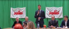 Czworo mężczyzn siedzących przy połączonych ławkach. Między nimi stoi z mikrofonem Minister Edukacji Narodowej – Dariusz Piontkowski. W tle zielony materiał i dwa transparenty z logo "Solidarność".