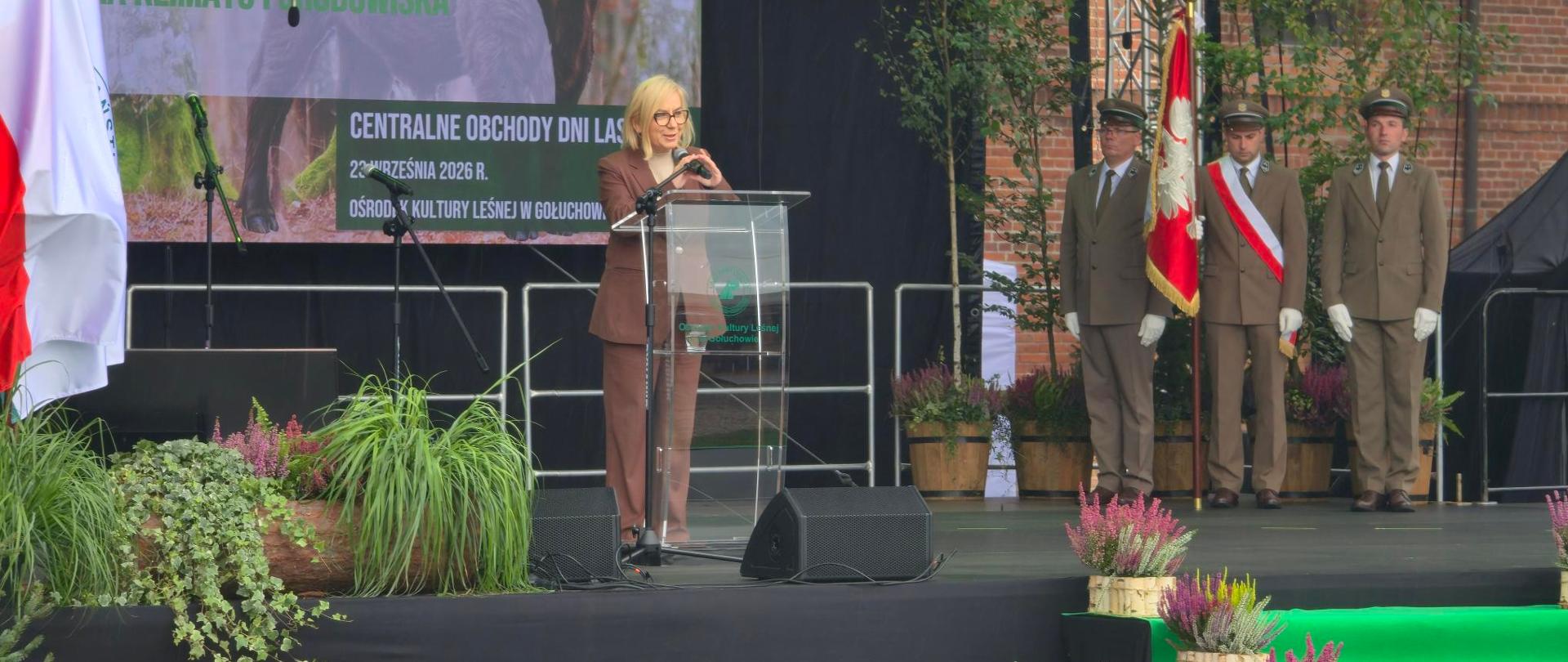 Ministra Paulina Hennig-Kloska podczas Centralnych Obchodów Dni Lasu. Ministra stoi za mównicą, za nią widoczny test telebim z prezentacją. Po prawej stronie zdjęcia stoją leśnicy ze sztandarem. Na pierwszym planie widoczne są elementy dekoracji sceny - kwiaty w doniczkach. 