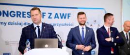 Minister Czarnek stoi za białą mównicą i mówi do mikrofonu, za nim ścianka z napisem Kongres WF z AWF, obok stoi kilku mężczyzn w garniturach.