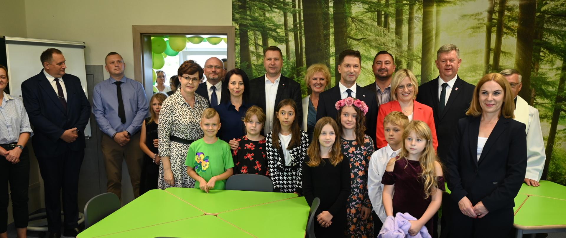 W szkolnej sali stoi duża grupa dzieci w kolorowych strojach, za nimi dorośli, wśród nich minister Czarnek, z przodu kilka zielonych stolików.