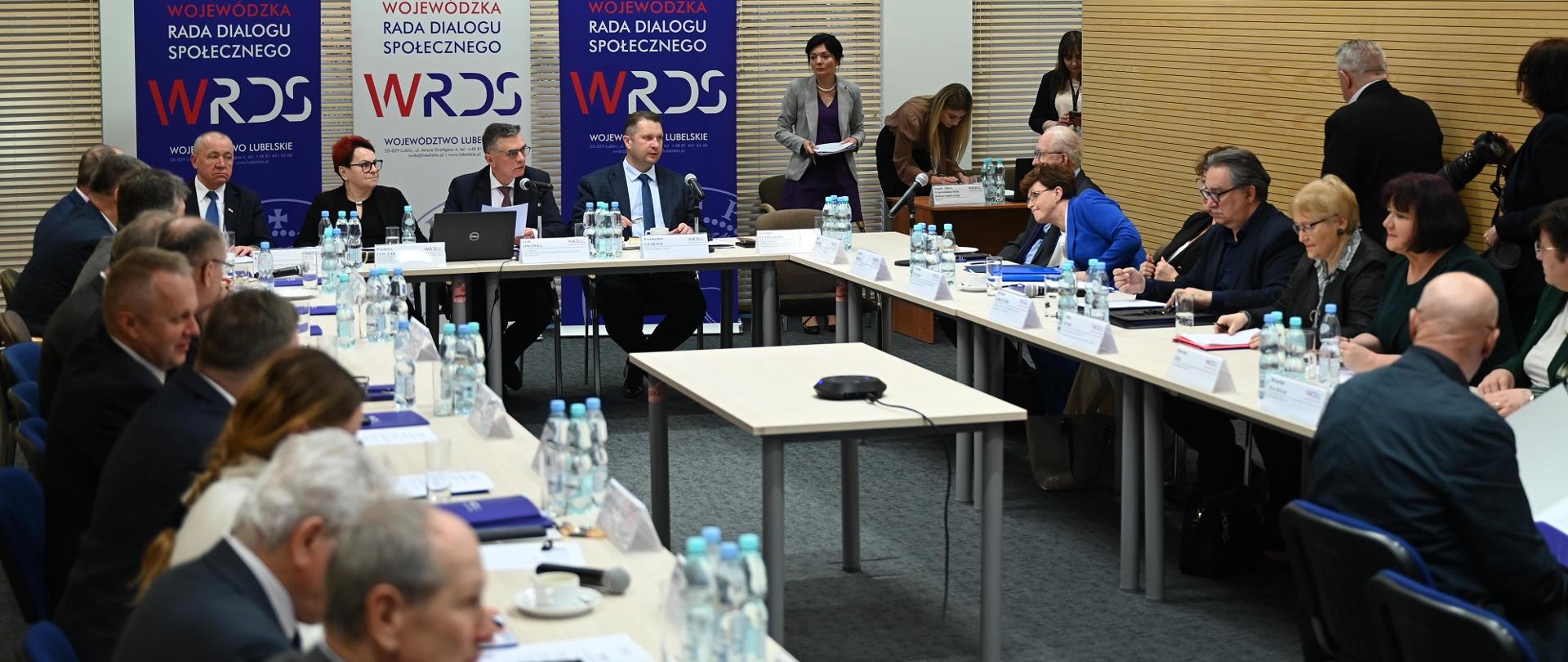 Widok na salę z żółtymi ścianami, przy ustawionych w czworokąt stołach siedzą ludzie, u szczytu stołu siedzi minister Czarnek, kobieta w czarnej marynarce i dwóch mężczyzn w garniturach, za nimi zasłonięte żaluzjami okna.