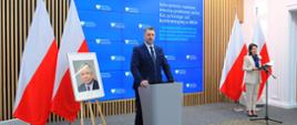 Minister Czarnek stoi za mównicą i mówi do dwóch mikrofonów, za nim niebieska ścianka z napisami Ministerstwo Edukacji i Nauki, obok portret Lecha Kaczyńskiego a za nim dwie polskie flagi, dalej za mikrofonem na stojaku stoi ubrana na biało kobieta, za nią kolejne dwie polskie flagi.