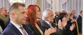 Widok z boku, na ustawionych w rzędy krzesłach siedzą ludzie, z przodu minister Czarnek i rudowłosa kobieta w czarnej marynarce, w tle zasłonięte białymi firankami okna.