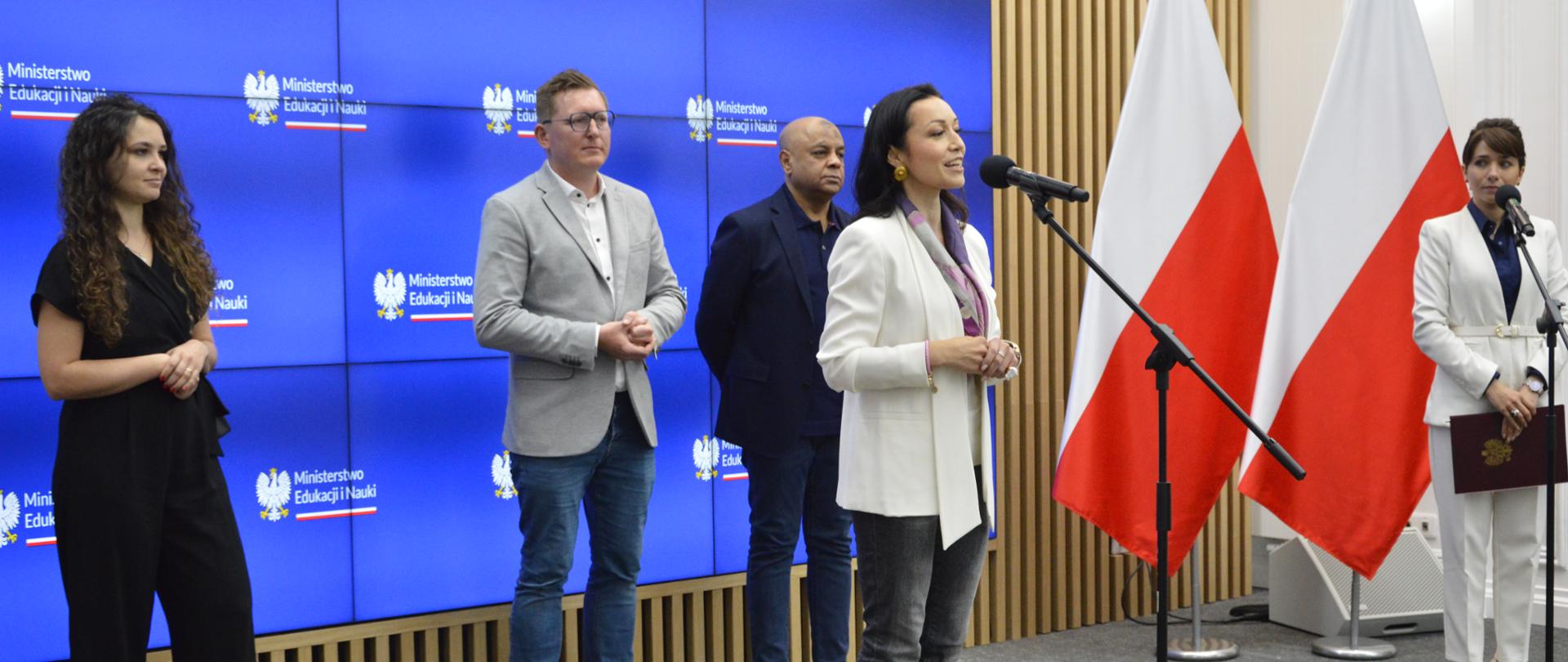 Na tle niebieskiej ścianki z napisem ministerstwo Edukacji i Nauki stoi mężczyzna w jasnoszarej marynarce, kobieta w czarnym ubraniu i mężczyzna w czarnej marynarce, przed nimi kobieta w białej bluzce mówi do mikrofonu na stojaku, dalej z boku dwie polskie flagi.