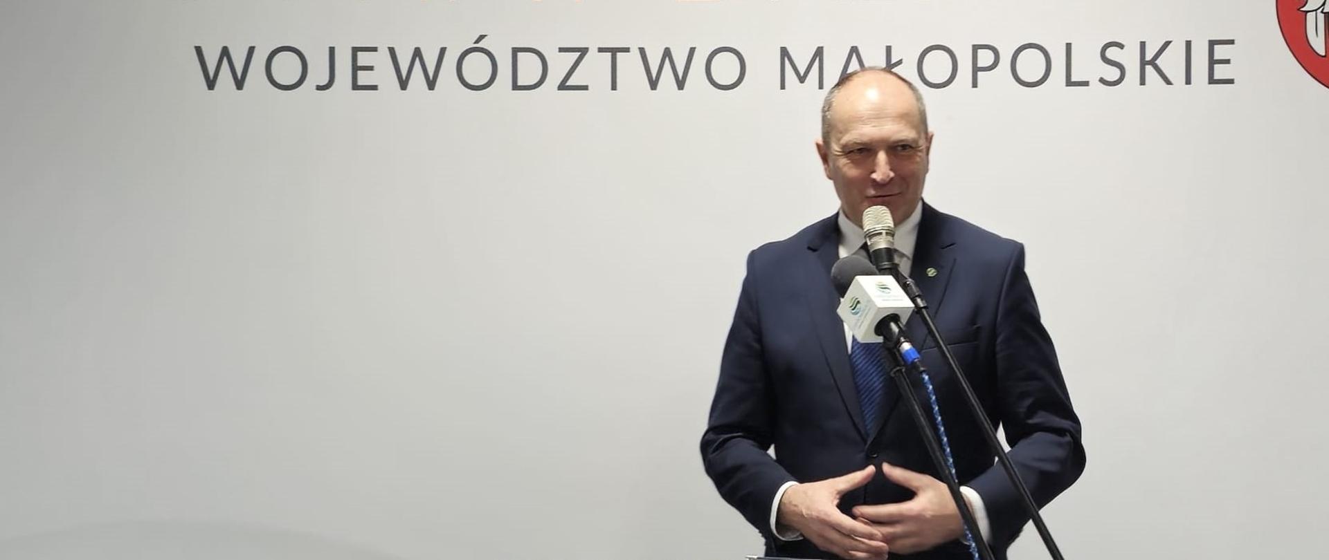 Wiceminister Henryk Kiepura przemawia. Minister stoi przy mównicy z herbem powiatu brzeskiego, a przed nim widoczne są mikrofony stacji telewizyjnych i radiowych.
