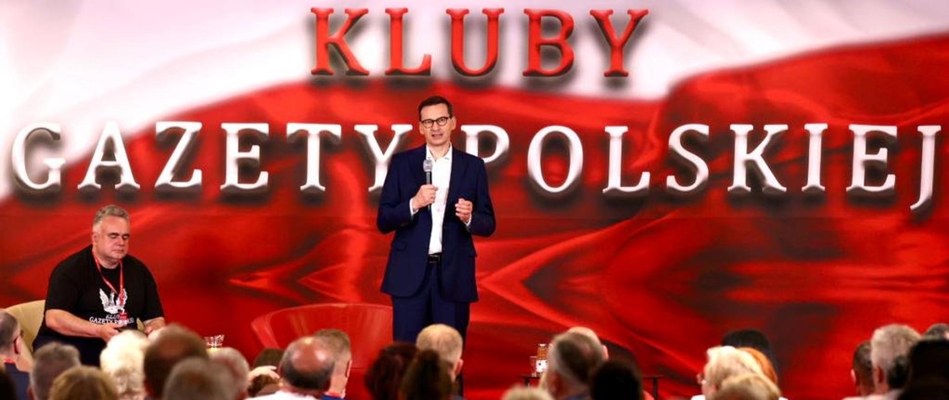 Na pierwszym planie tłum siedzących ludzi. Premier Mateusz Morawiecki na scenie. W tle napis: Kluby Gazety Polskiej. 