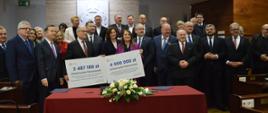 Duża grupa ludzi stoi przy czerwonym stole, kilka osób z przodu trzyma dwa duże symboliczne czeki z napisami 3 487180 zł i 4 000 000 zł.