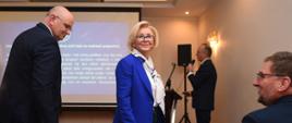 Widok z tyłu na przód sali, wiceminister Machałek w niebieskim ubraniu idzie wzdłuż rzędu krzeseł, za nią mężczyzna w czarnym garniturze, dalej widać ekran z napisem Rodzina w centrum pracy wychowawczej szkoły i mężczyznę mówiącego do mikrofonu.