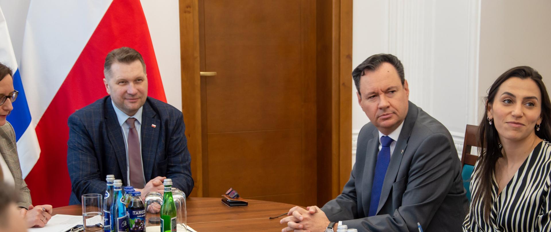Za drewnianym stołem siedzi minister Czarnek, obok niego mężczyzna w szarym garniturze i niebieskim krawacie i kobieta w bluzce w pionowe biało-czarne paski, za nimi flagi Polski i Izraela, dalej biała ściana i drewniane drzwi.