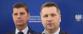 Minister Czarnek mówi do mikrofonu, za nim stoi wiceminister Piontkowski, dalej niebieska ściana.