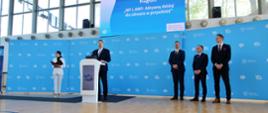 Na sali gimnastycznej z drewnianą podłogą stoi minister Czarnek za białą mównicą i mówi do mikrofonu, obok niego stoi kobieta w białym garniturze, z tyłu trzech mężczyzn w garniturach, za nimi niebieska ściana i duże okna.
