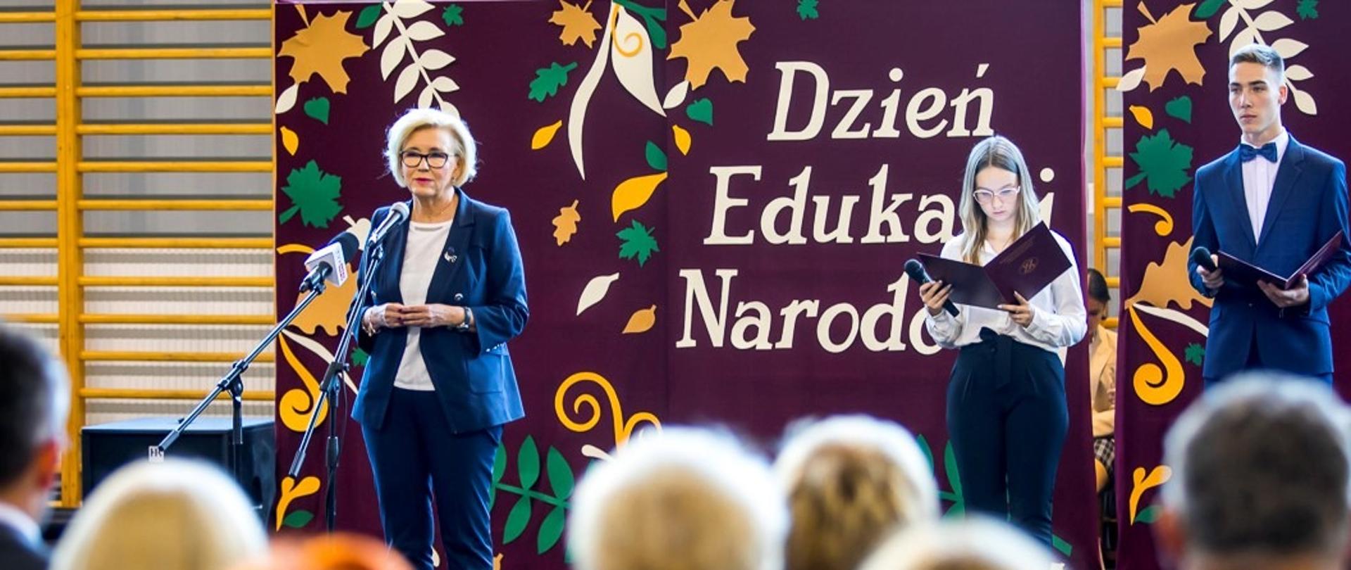 Wiceminister Machałek stoi na scenie, obok stoi uczeń i uczennica, tyłem widać siedzących nauczycieli
