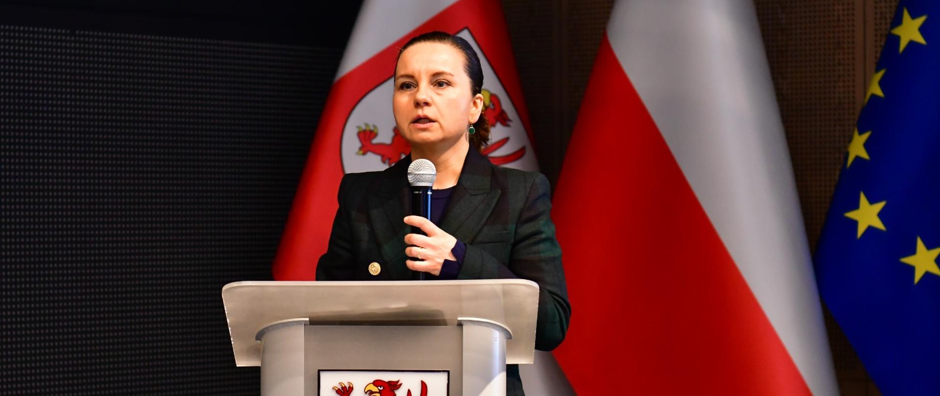 Na zdjęciu Wiceministra Urszula Zielińska podczas konferencji Wojewódzkiego Funduszu Ochrony Środowiska w Szczecinie. Stoi za mównicą na której widoczny jest herb województwa zachodniopomorskiego. Za nią widoczne są trzy flagi - flaga województwa zachodniopomorskiego, flaga Polski i flaga Unii Europejskiej. 