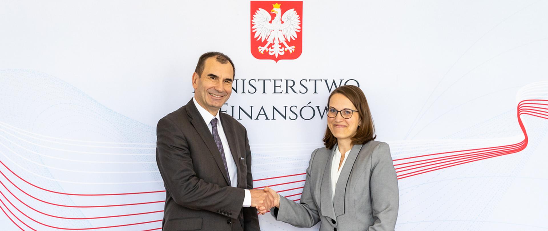 Minister Magdalena Rzeczkowska i Allain Pilloux Wiceprezes Europejskiego Banku Odbudowy i Rozwoju podają sobie dłonie na tle banera Ministerstwa Finansów
