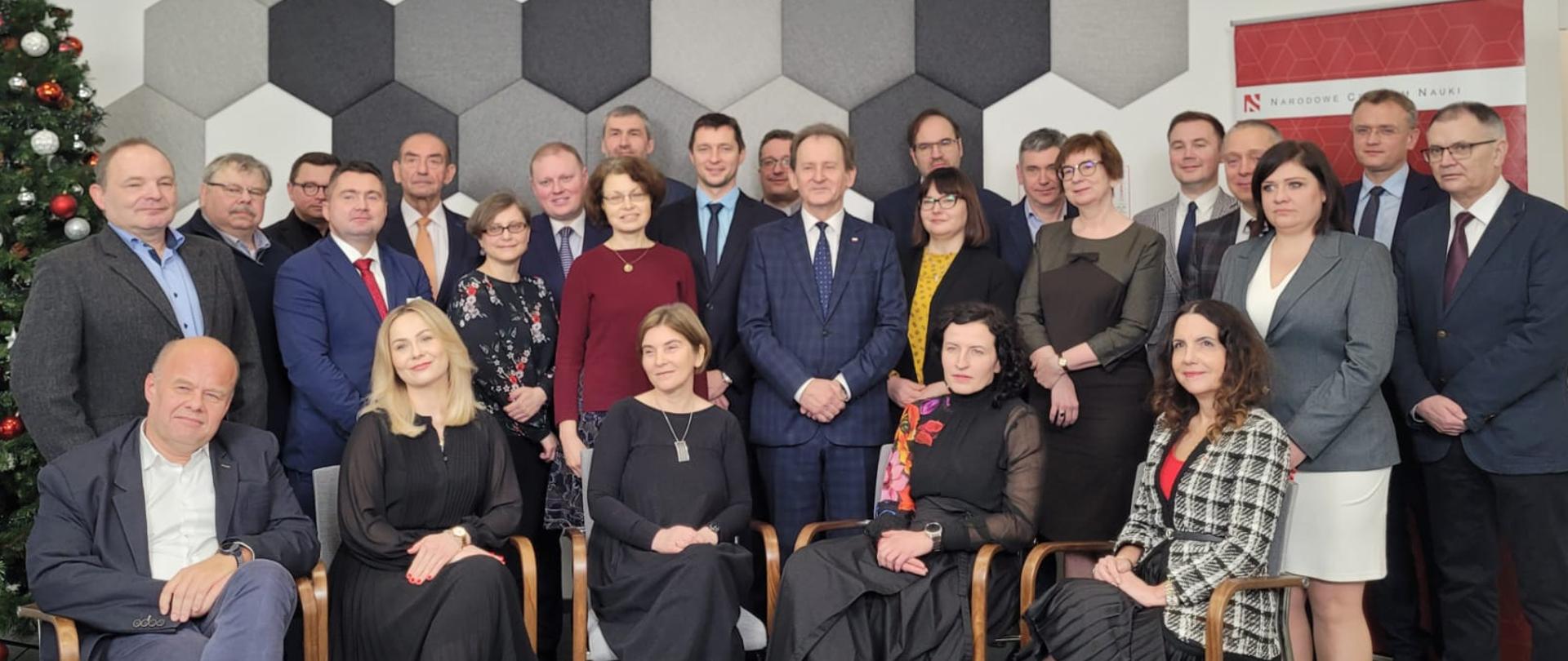 Zdjęcie zbiorowe, uczestnicy uroczystości stoją i siedzą na krzesłach, za nimi z lewej choinka z bombkami, dalej ściana we wzór z sześciokątów o różnych odcieniach szarości.