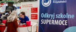 Duża hala, z przodu niebieski baner z napisem Laboratoria przyszłości - odkryj szkolne supermoce, za nim białe stoisko na którym kobieta w niebieskiej koszulce pokazuje coś kobiecie w czerwonym płaszczu.