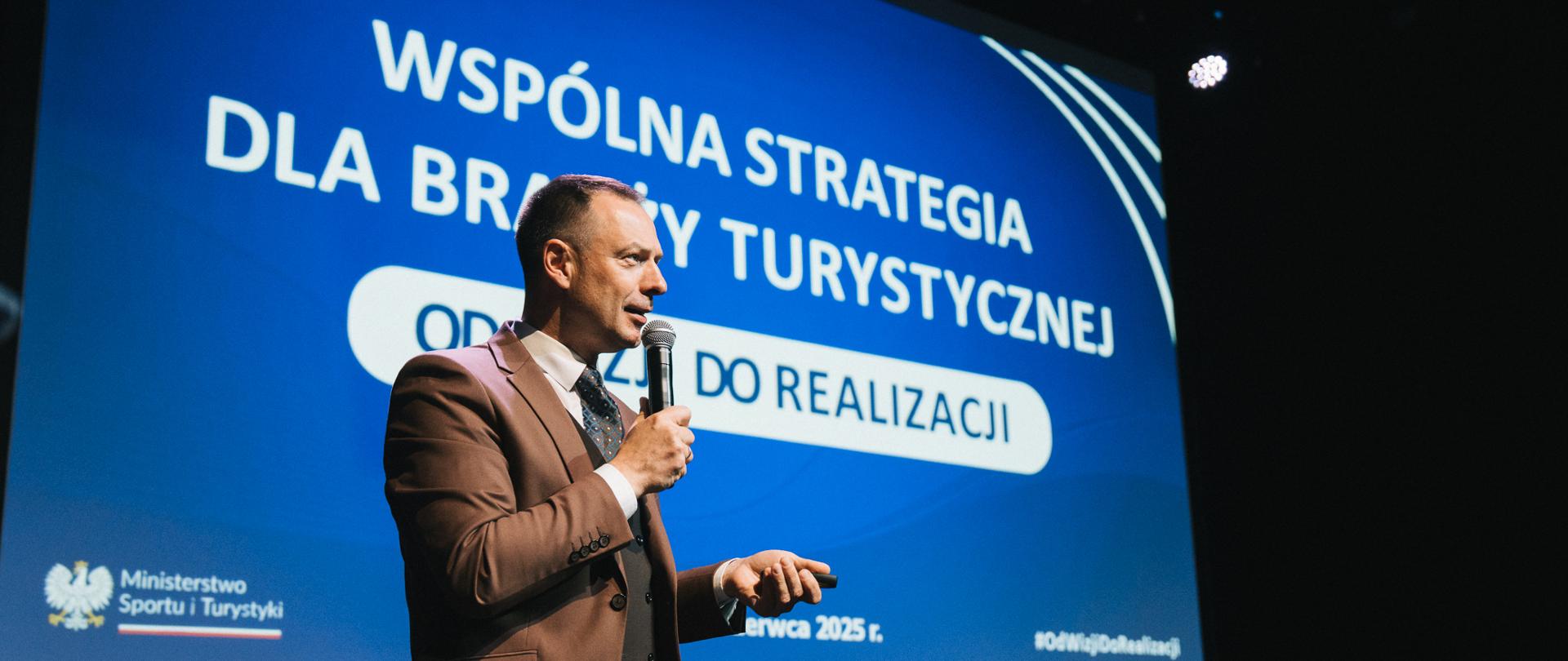 Piotr Borys na konferencji w Gdańsku na tle niebieskiego ekranu z prezentacją z danymi