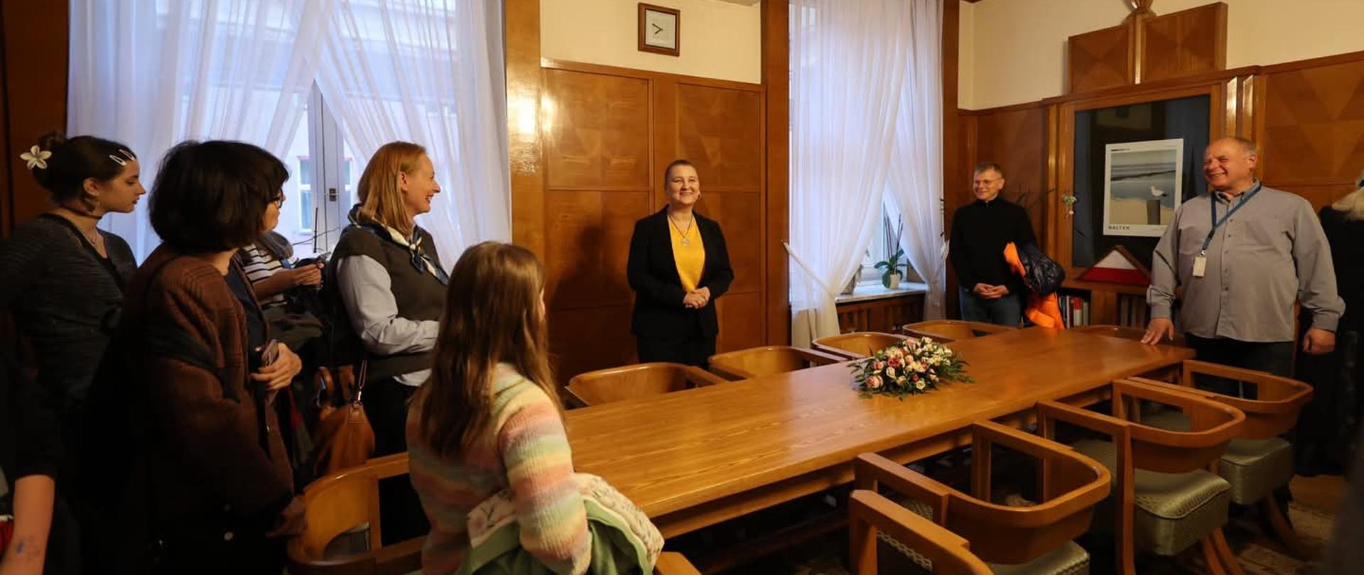 Wnętrze pomieszczenia z drewnianymi panelami na ścianach i dużym drewnianym stołem owalnym pośrodku. Przy stole stoi grupa osób, skierowanych w stronę kobiety stojącej za stołem, po jego krótszej stronie. Kobieta ma blond włosy i ubrana jest w czarną marynarkę i żółtą bluzkę. Uśmiecha się i wydaje się mówić do zgromadzonych.
Po lewej stronie stołu stoi kilka osób, częściowo zasłoniętych, w różnym wieku i ubranych w codzienne stroje. Po prawej stronie stołu stoi dwóch mężczyzn. Jeden z nich jest łysy i ubrany w szarą koszulę z długim rękawem i niebieską smycz na szyi. Drugi mężczyzna, stojący za nim, ma siwe włosy i ubrany jest w czarną bluzę.
Na stole leży bukiet różowych i białych kwiatów. W tle widoczne są okna z białymi, przezroczystymi zasłonami, klimatyzator na ścianie oraz drzwi z plakatem. Oświetlenie jest jasne, sufitowe. Atmosfera sugeruje spotkanie lub prezentację w kameralnym gronie.