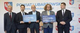 Pod ścianką z napisem Powiat Lubartowski stoi minister Czarnek i czterech mężczyzn w garniturach, dwóch z nich trzyma tabliczki z napisami Powiat lubartowski.