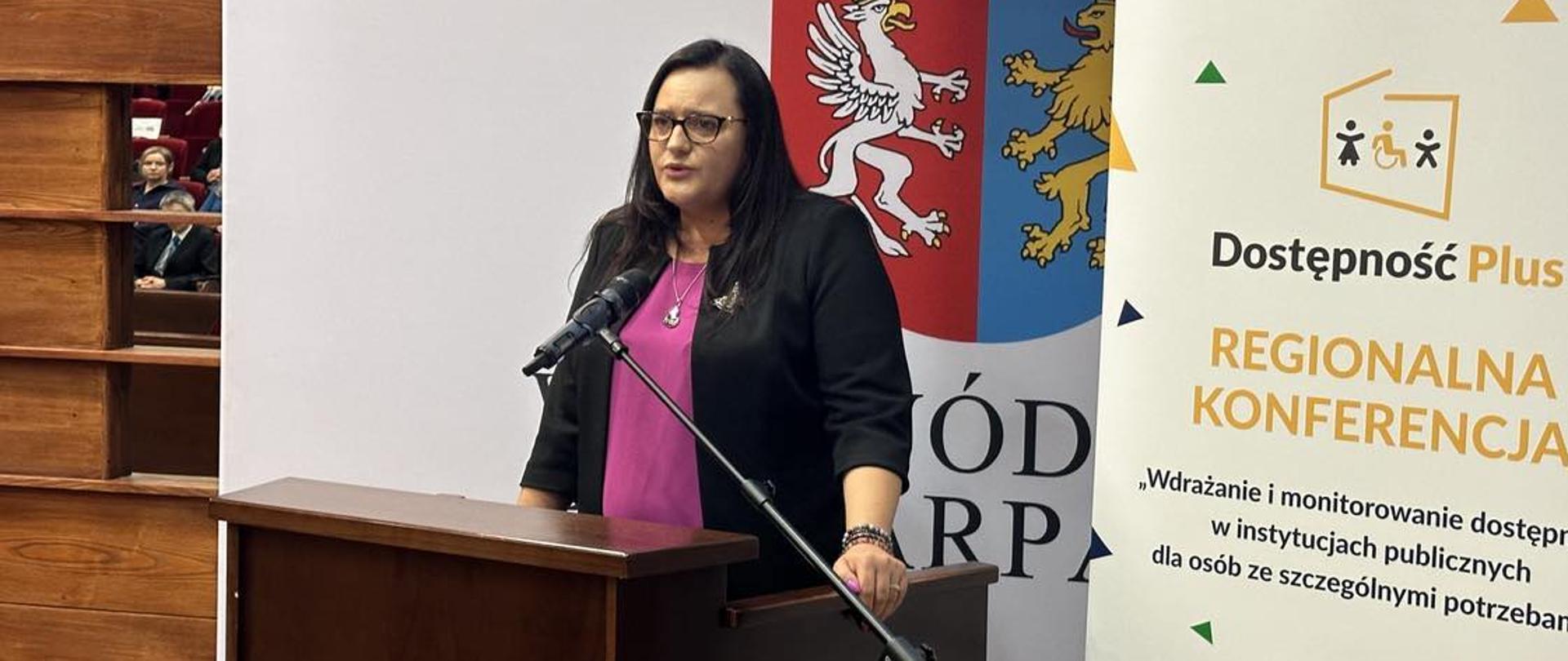 Wiceminister Małgorzata Jarosińska-Jedynak w mównicy z mikrofonem na tle banerów