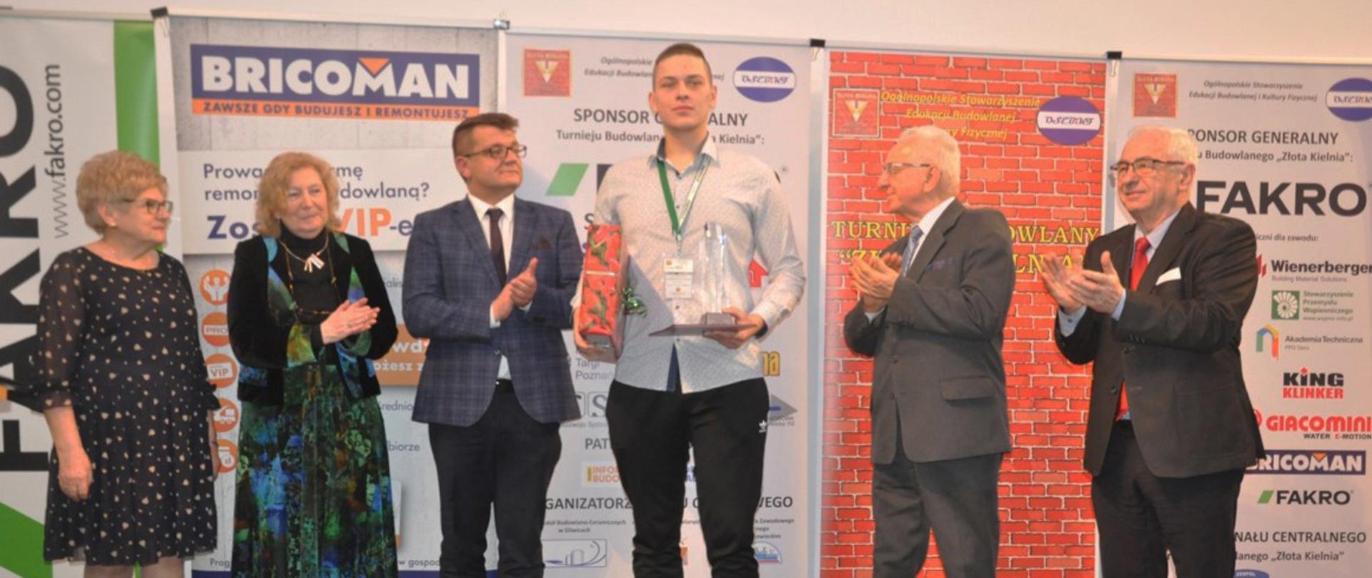 Laureat konkursu odbiera dyplom i statuetkę. Za nim widać stojące reklamy organizatorów konkursu. 