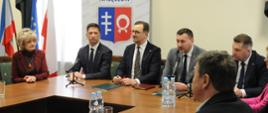 Przy ustawionych w czworokąt drewnianych stołach siedzi kilka osób, za nimi ścianka z niebiesko-czerwonym herbem Nałęczowa, za nią żółta ściana, z lewej przysłonięte zasłonami okno.