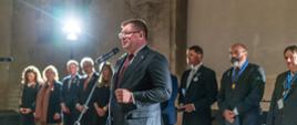 W oświetlonej jaskrawymi reflektorami sali z kamiennymi ścianami stoi wiceminister Rzymkowski i mówi do mikrofonu na stojaku, za nim stoi szereg ludzi.