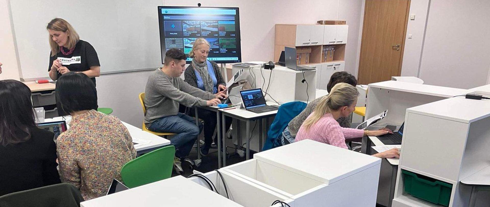Widok na salę, przy białych meblach przy laptopach siedzi kilka osób, na ścianie wisi duży monitor.