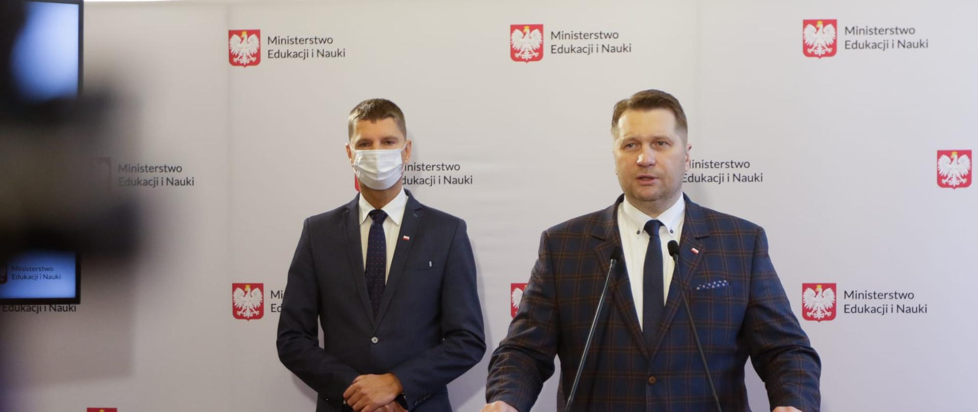 Podczas konferencji minister Przemysław Czarnek przemawia do mikrofonu. Z lewej strony stoi minister Piontkowski. Na tle białej ścianki z napisem ministerstwo edukacji i Nauki