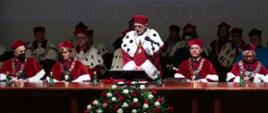 Za drewnianym stołem siedzi kilkanaście osób w ceremonialnych uniwersyteckich czerwonych strojach, mężczyzna na środku stoi i mówi do mikrofonu, przed nim na podłodze bukiet biało-czerwonych kwiatów.